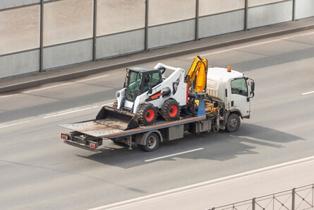White tow truck carrying a small white construction vehicle on a road